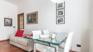 Ein modernes Wohnzimmer mit einem Glastisch, weißen Stühlen, einem grauen Sofa mit roten und gemusterten Kissen sowie einem gerahmten Gemälde an der Wand.
