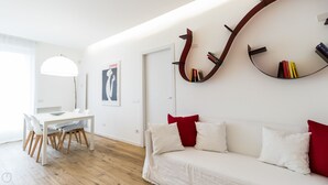 En moderne stue med en hvid sofa, røde puder, et træspisebord med stole og et vægmonteret bogreol.