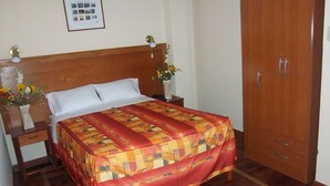 A neatly made bed with a patterned bedspread, a wooden headboard, a nightstand with a vase of flowers, and a wooden wardrobe. - Hotel Moquegua (Moquegua)