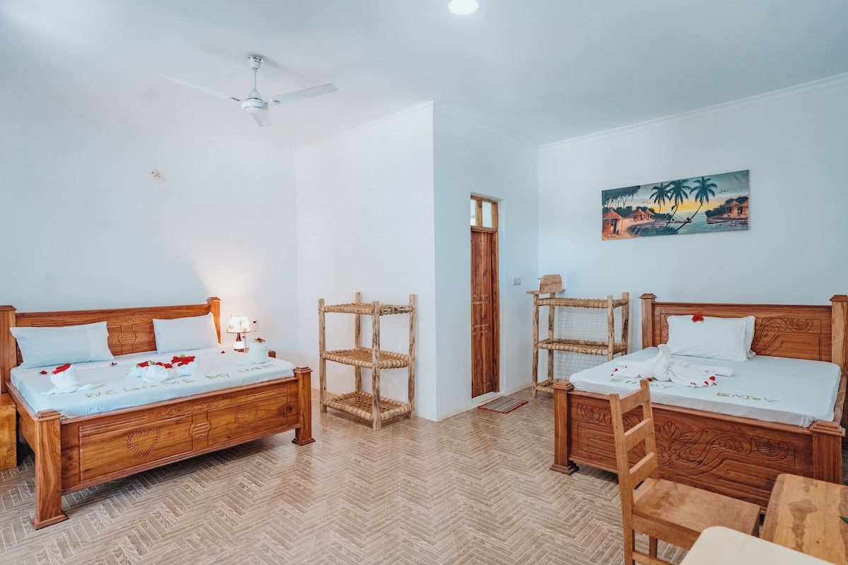 Ein Zimmer mit zwei Holzbett, einem Deckenventilator, einem Holzstuhl, einem gewobenen Regal und einem Bild an der Wand.