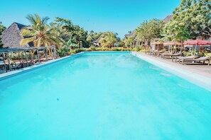 Une piscine d’un bleu éclatant, entourée de palmiers et de petites huttes aux toits de chaume, avec des chaises longues et des parasols sur le côté droit.