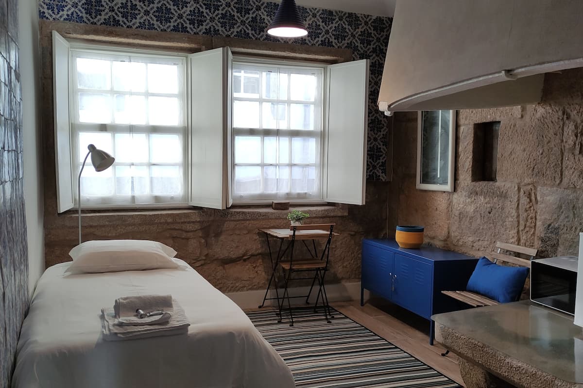 A bedroom with a bed, a desk, a chair, a window, and a stone wall.