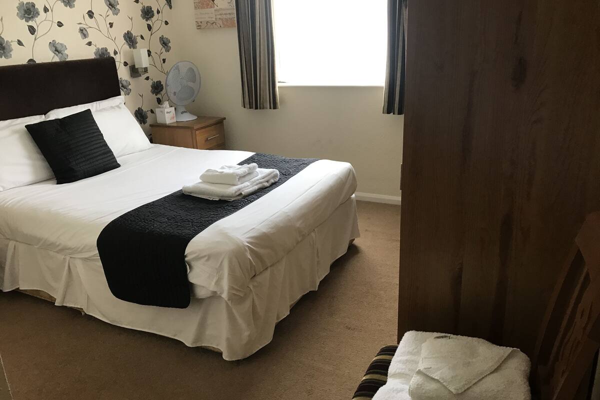 A neatly made bed with white and black bedding, a bedside table with a lamp, and a floral wallpaper pattern.