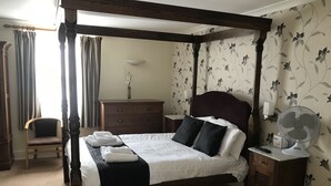 A four-poster bed with a dark headboard, a wooden nightstand with a fan, a dresser, and a chair in a room with floral wallpaper.