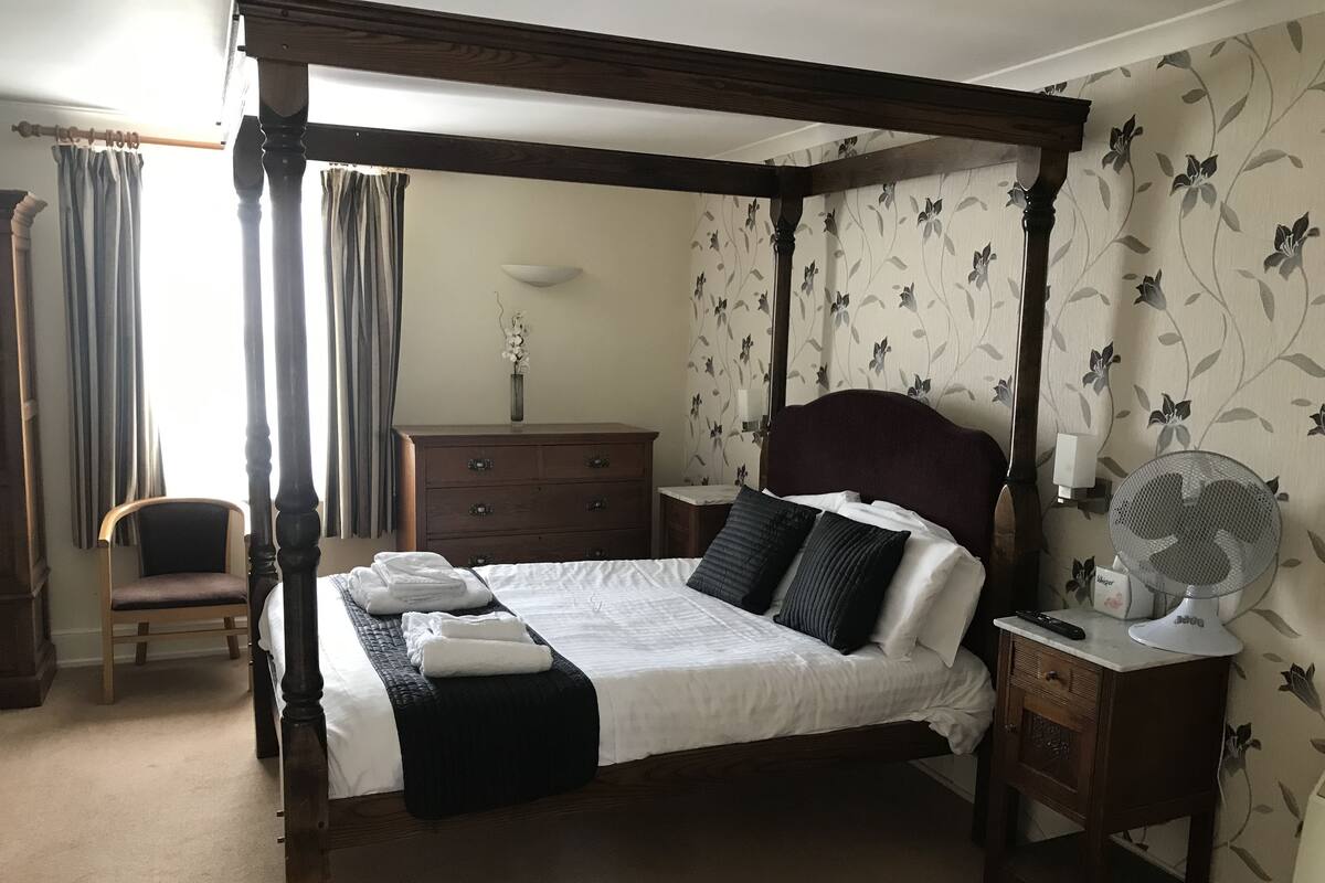 A four-poster bed with a dark headboard, a wooden nightstand with a fan, a dresser, and a chair in a room with floral wallpaper.