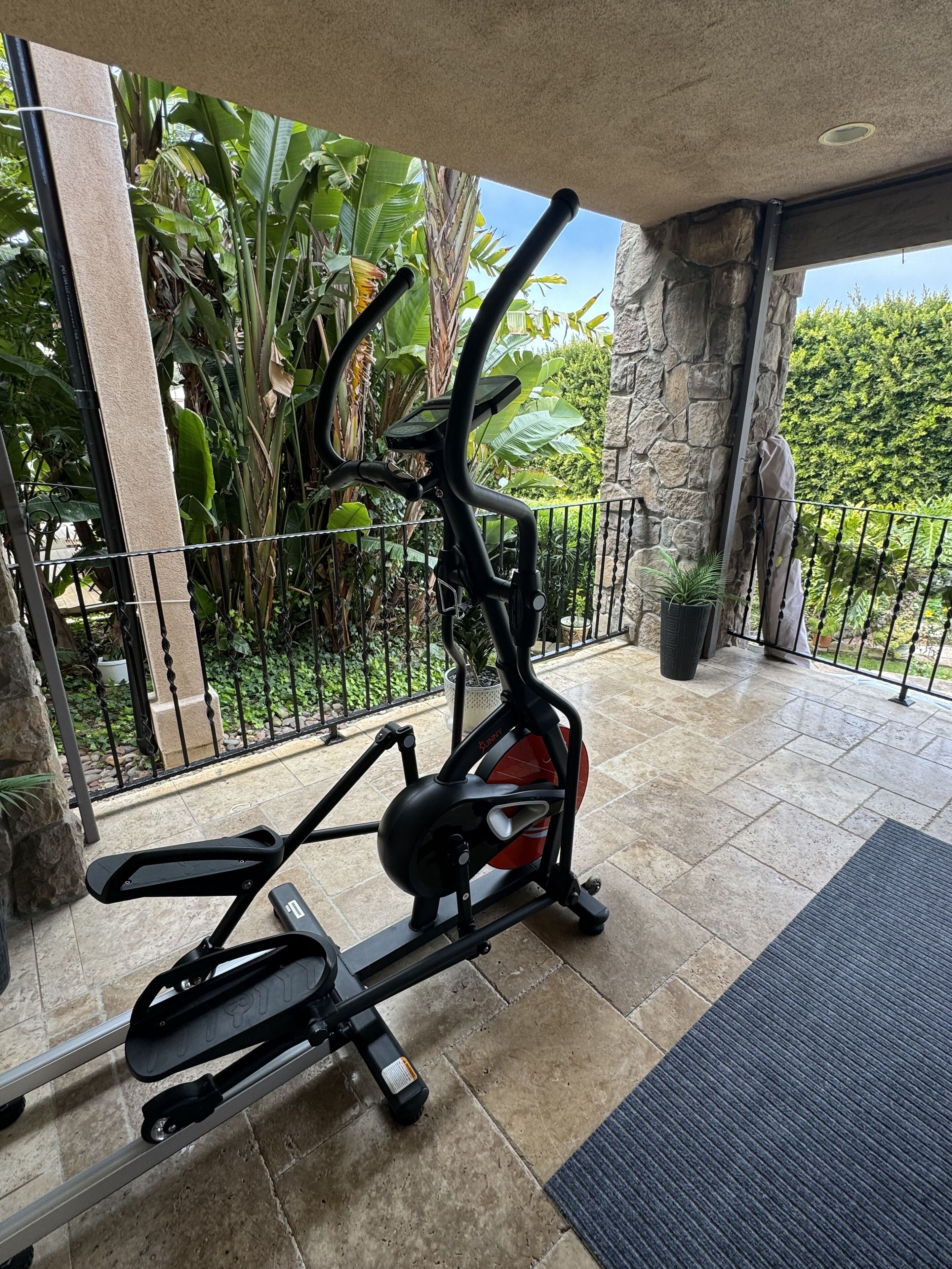 An outdoor exercise area with an elliptical machine, surrounded by a stone wall and lush greenery.
