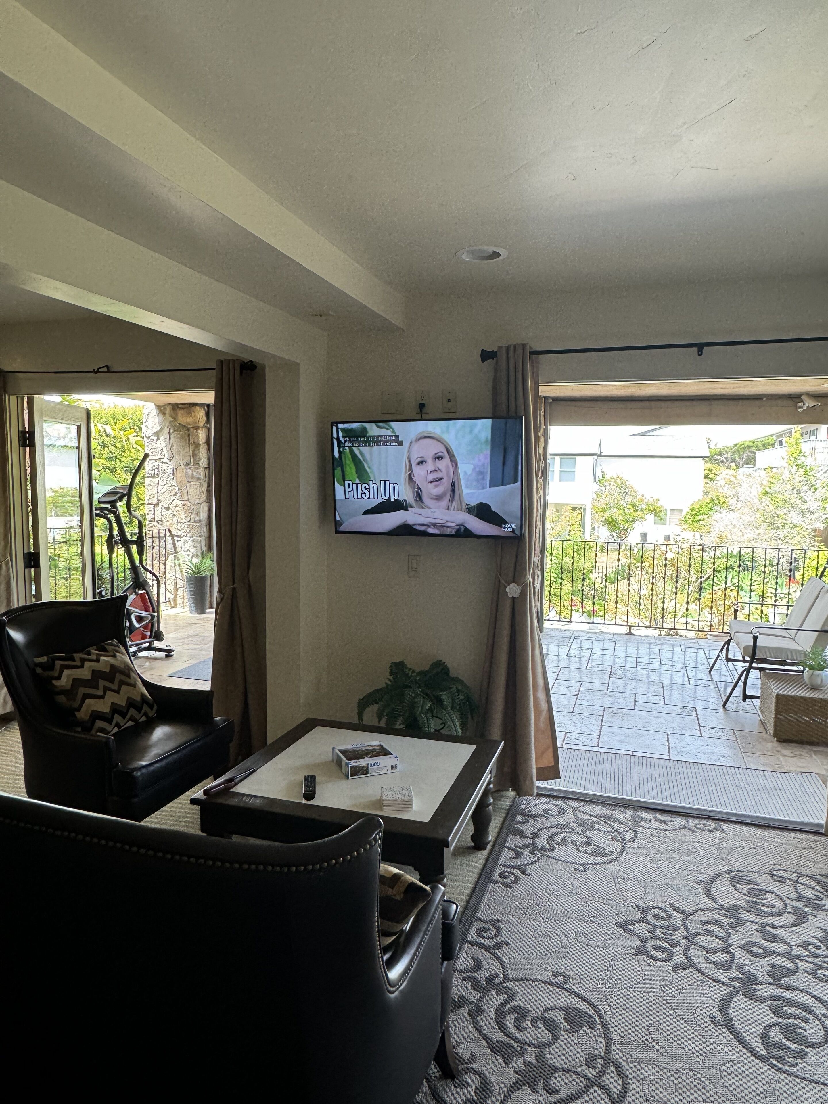 A living room with a television mounted on the wall, a black leather sofa, a coffee table, and a view of the outdoor patio area.