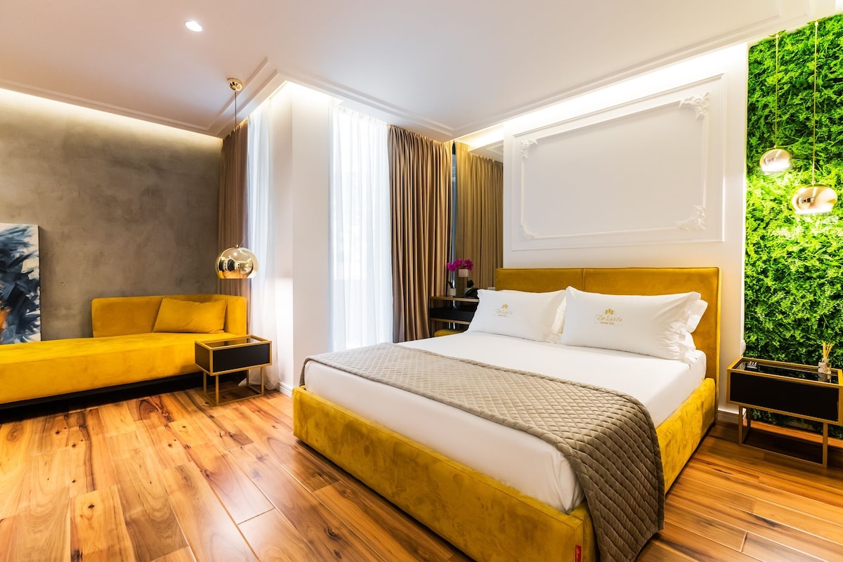 Une chambre d’hôtel moderne avec un plancher en bois, un canapé jaune, un lit avec une literie blanche et un mur vert.