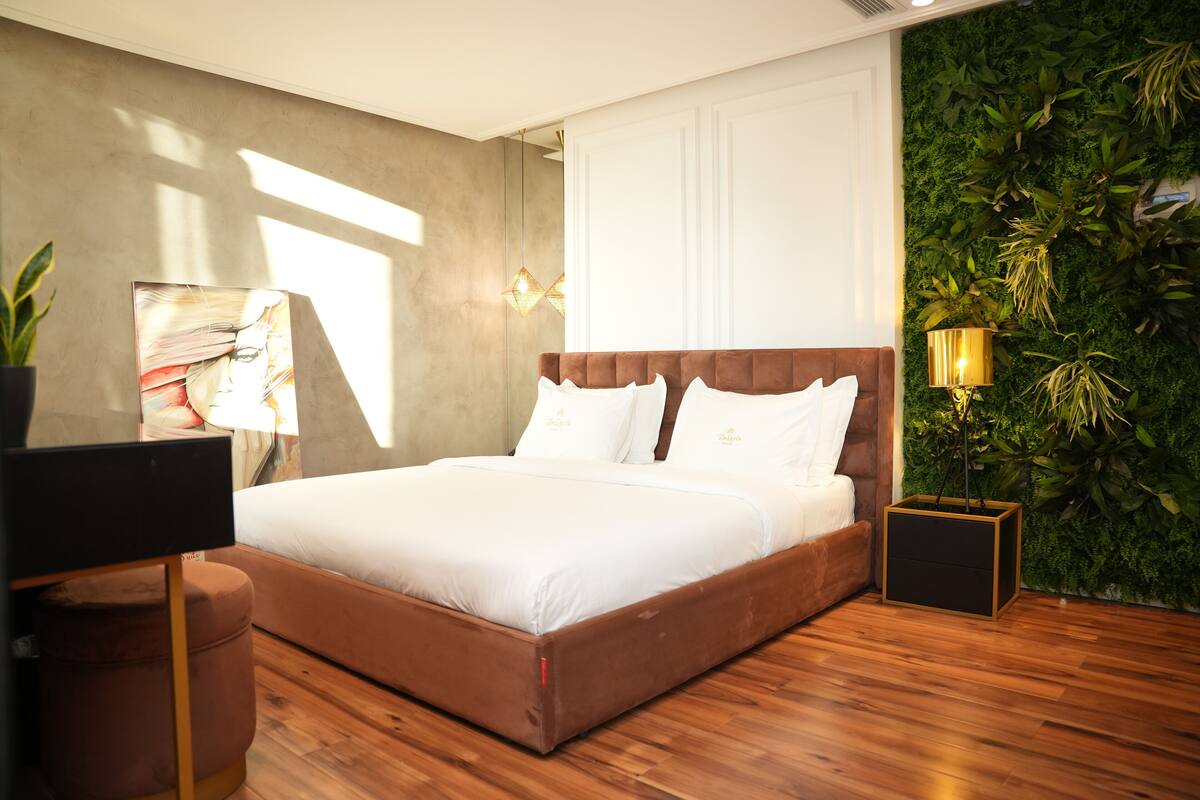 Une chambre moderne avec un plancher en bois, un lit recouvert d’un tissu marron, du linge de lit blanc, un grand tableau, une plante en pot et un mur vert orné de plantes.