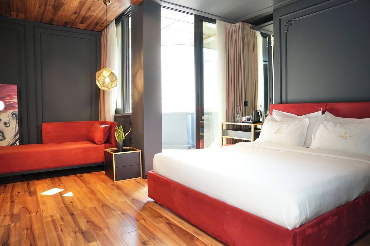 Une chambre d’hôtel moderne avec un canapé rouge, un grand lit avec du linge de lit blanc, un plancher en bois et une grande fenêtre avec des rideaux.