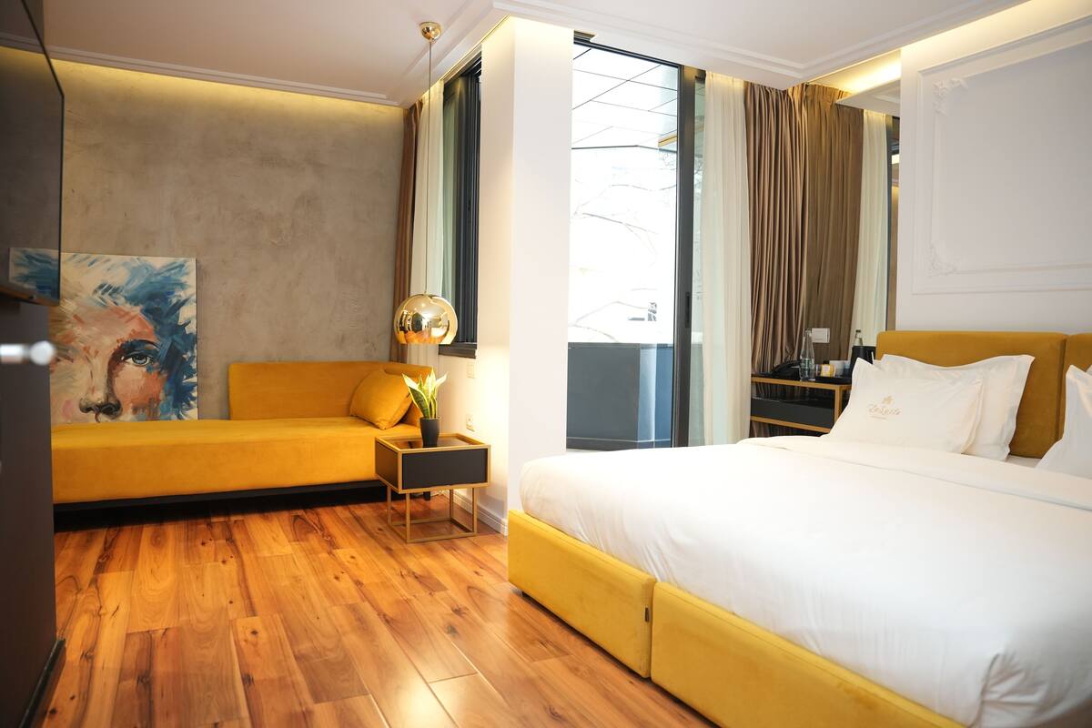 Une chambre d’hôtel moderne avec un plancher en bois, un canapé jaune, un grand lit avec une literie blanche, un tableau au mur et une vue sur l’extérieur par une fenêtre.
