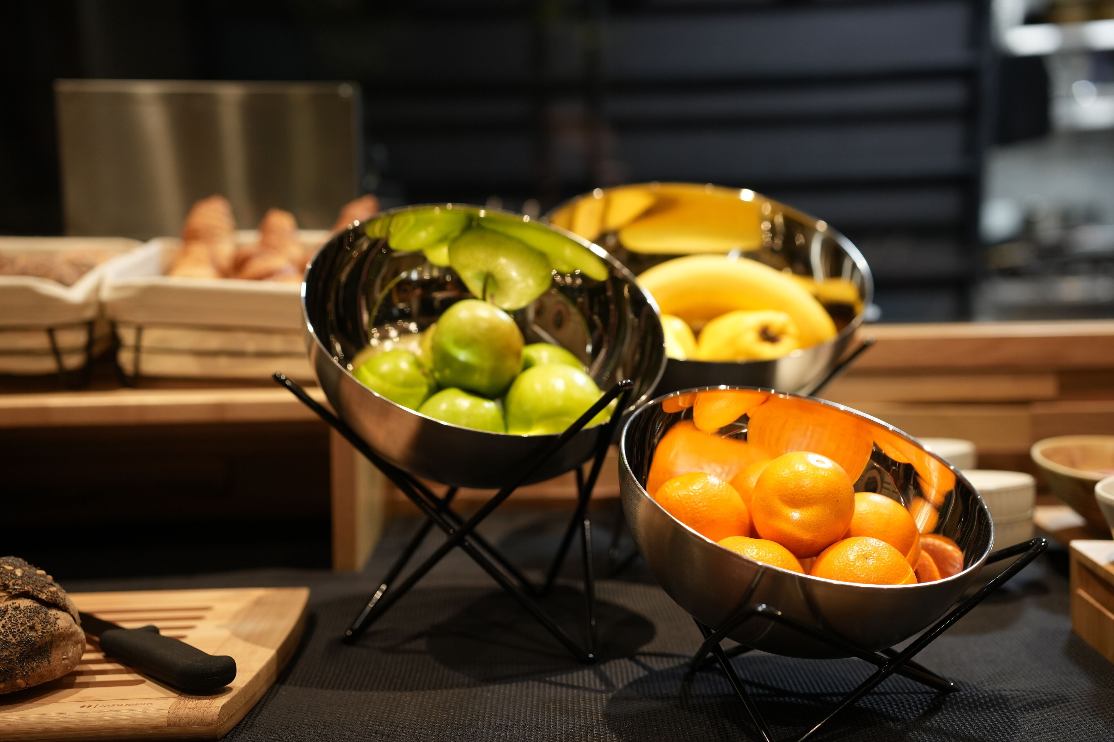 A kitchen counter with three metal bowls containing fruits, a cutting board with a knife, and a loaf of bread.