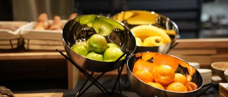 A kitchen counter with three metal bowls containing fruits, a cutting board with a knife, and a loaf of bread.