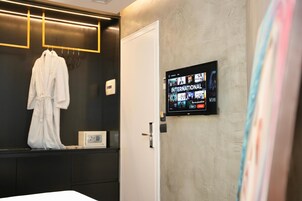 Habitación de hotel con un armario de color oscuro, una bata blanca colgada, un televisor mostrando opciones de películas y una puerta con manija.