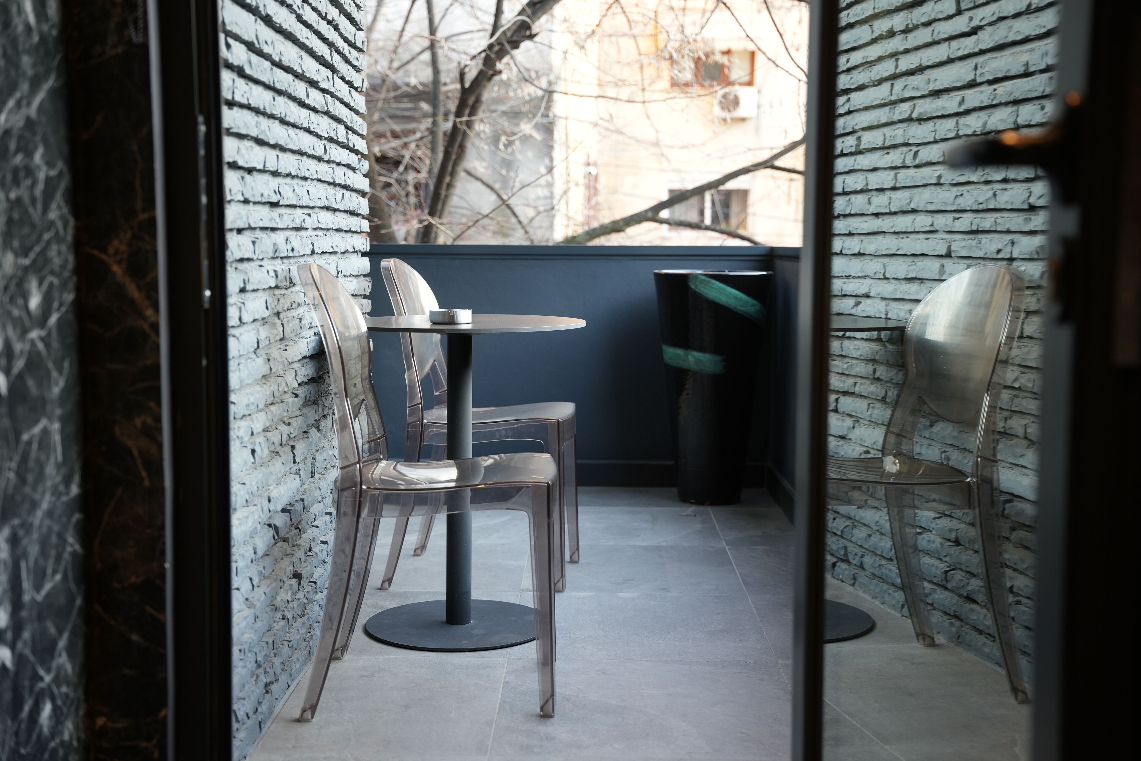 Un espacio al aire libre moderno con una mesa redonda y sillas, un cubo de basura y vistas a una pared de ladrillo y ramas de árboles.