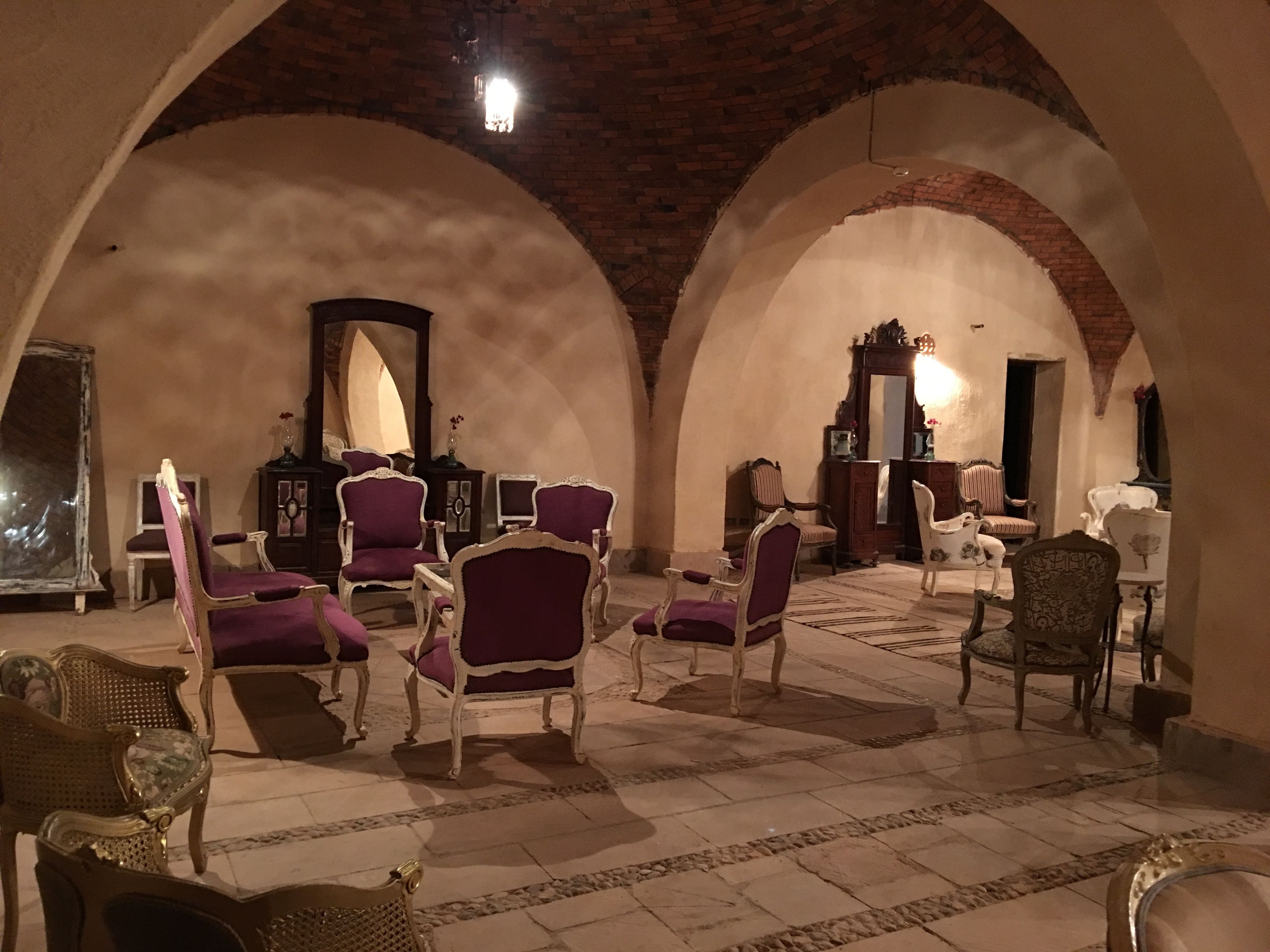 A room with arched ceilings and a tiled floor, featuring several purple and white upholstered chairs and a wooden cabinet with a mirror.