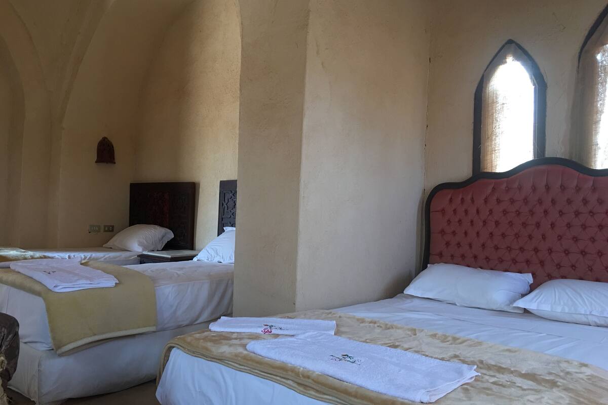 A hotel room with two beds, each with white linens and a brown blanket. There are two windows with arched tops, and the room has a vaulted ceiling.