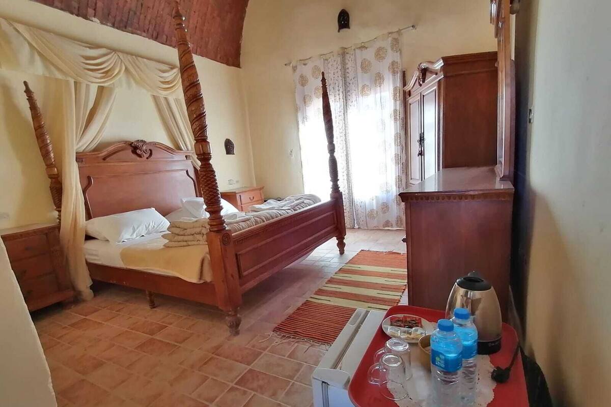 A bedroom with a four-poster bed, a wooden nightstand, a dresser, and a small table with a kettle and water bottles.