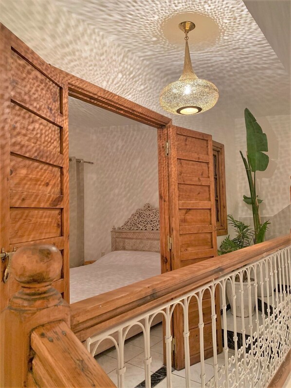 Une chambre avec une porte en bois, un lit, un luminaire suspendu et une rambarde de balcon.