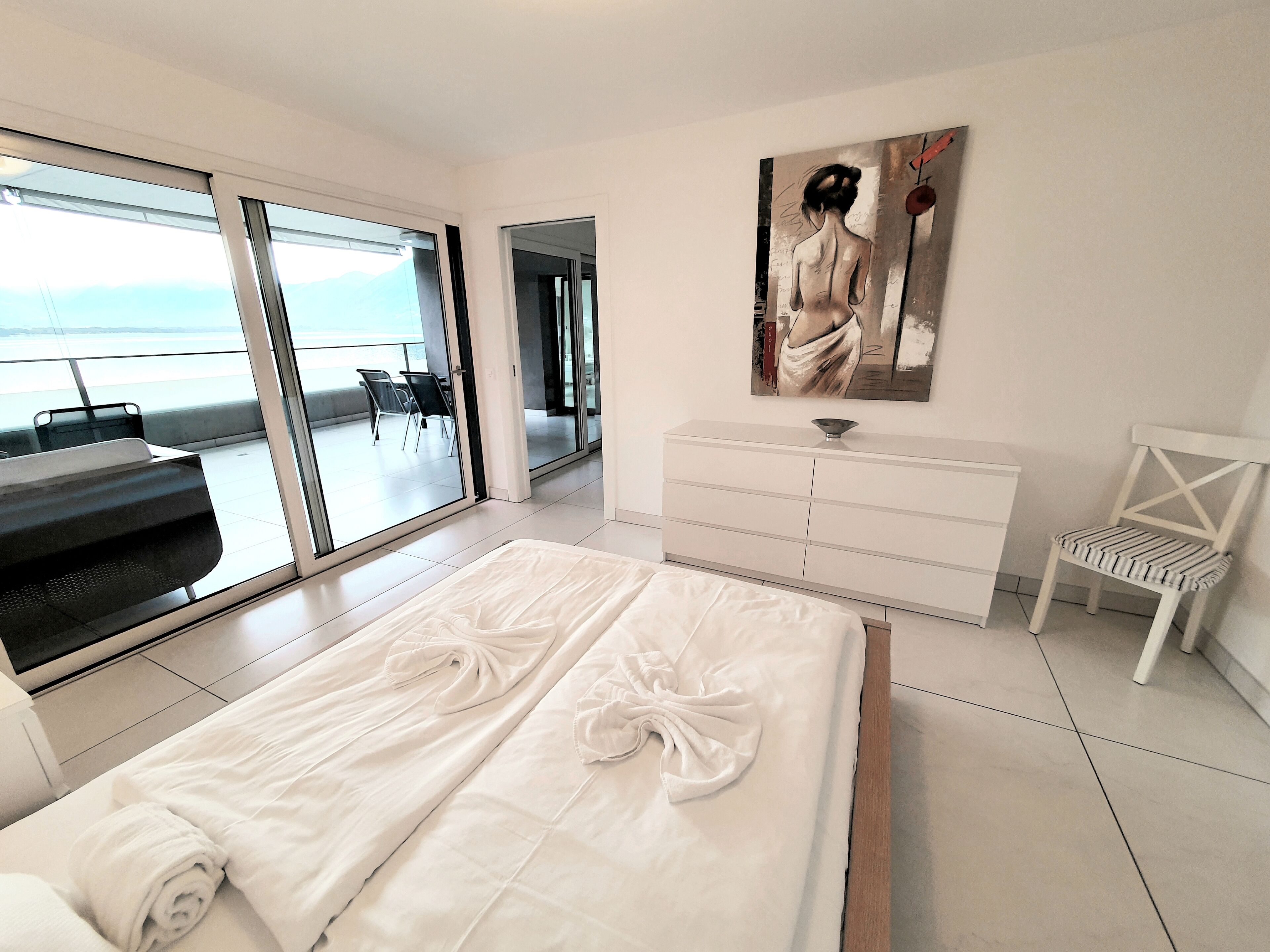 A modern hotel room with a large bed, a white dresser, a chair, and a painting on the wall.
