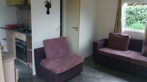 Un espace de vie compact comprenant un coin cuisine, un fauteuil violet, un canapé et une fenêtre avec des rideaux.