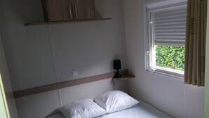 Une petite chambre à coucher, bien rangée, avec un lit simple, une table de chevet avec une lampe, une étagère au-dessus du lit et une fenêtre avec des rideaux.
