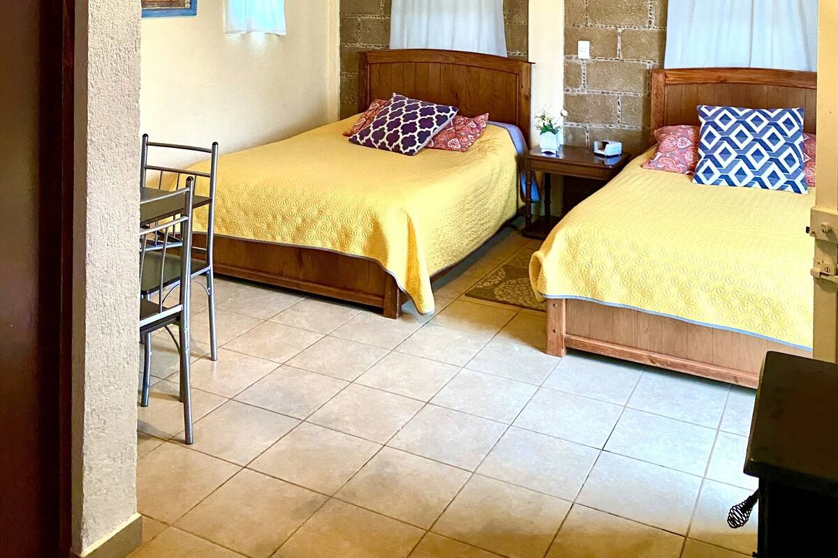 A room with two beds, wooden ceiling beams, a chandelier, a small table, and a chair.