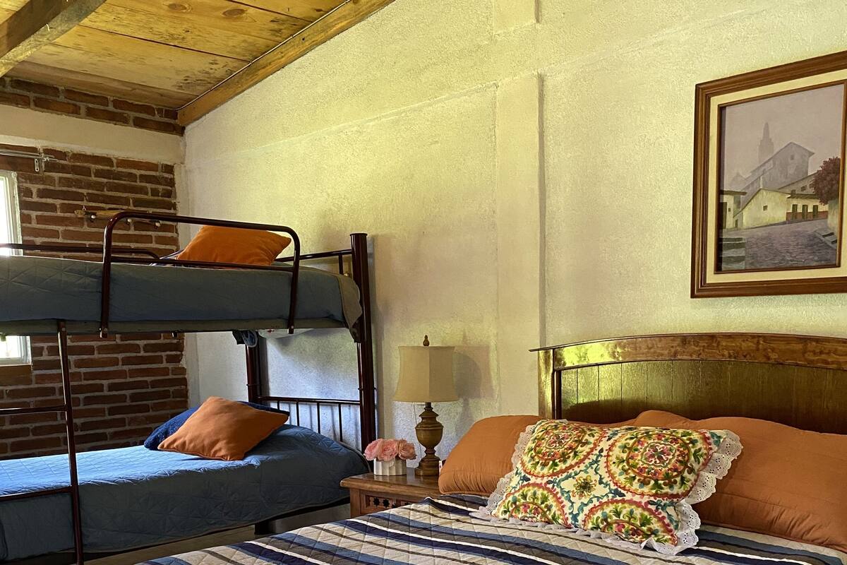 A room with a bunk bed, a single bed with a striped quilt, a nightstand with a lamp, and a framed picture on the wall.