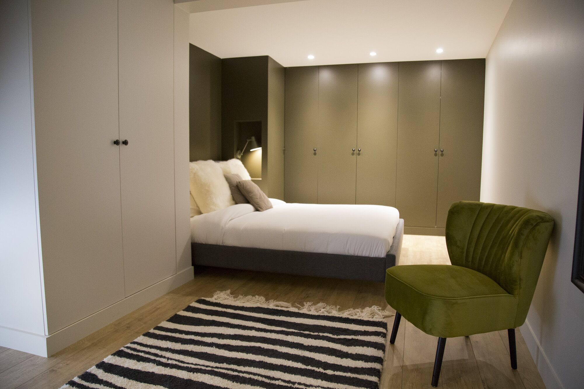 A bedroom with a bed, a green armchair, a striped rug, and a wardrobe.