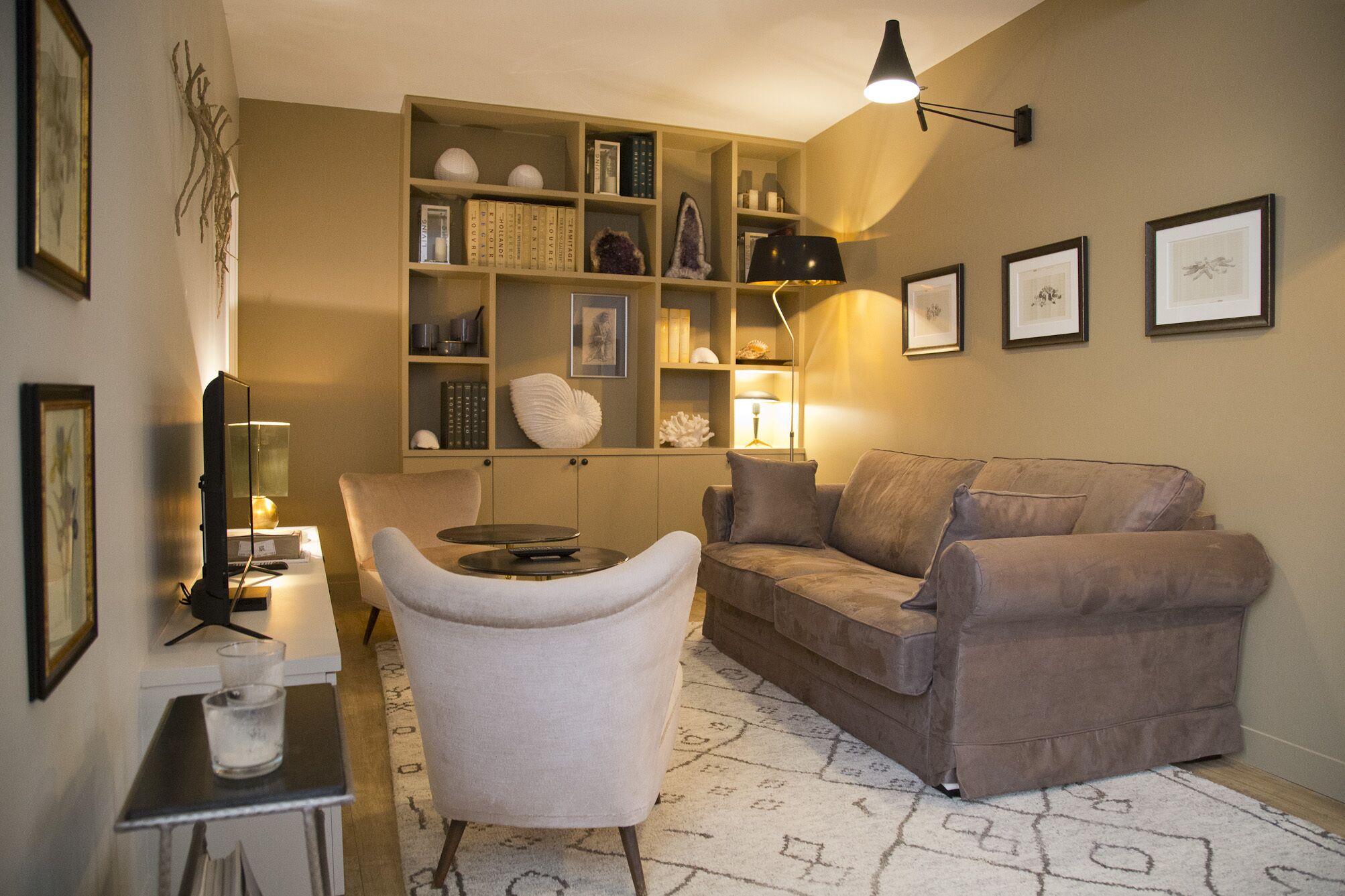 A cozy living room with a beige sofa, a white armchair, a small table with a lamp, and a bookshelf filled with books and decorative items.