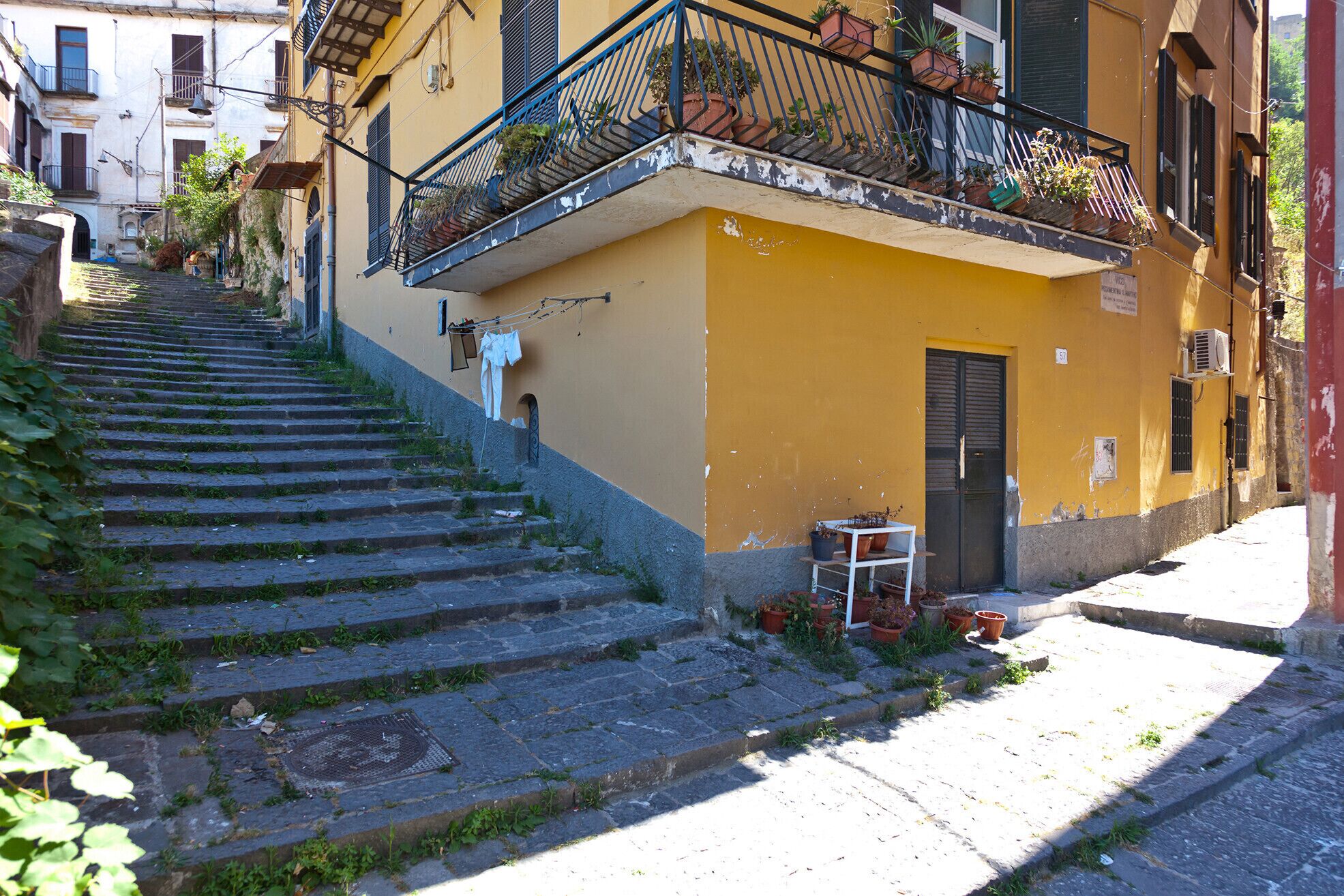 A narrow street with a yellow building, a staircase leading up, and potted plants on the balcony.