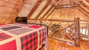 Une chambre mansardée chaleureuse avec un plafond en bois, un lit recouvert d’une couette à carreaux rouges et noirs, et une rambarde en bois rustique.