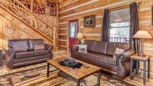 L’intérieur d’une cabane en rondins, avec deux canapés en cuir, une table basse en bois, un escalier et une fenêtre avec des rideaux.