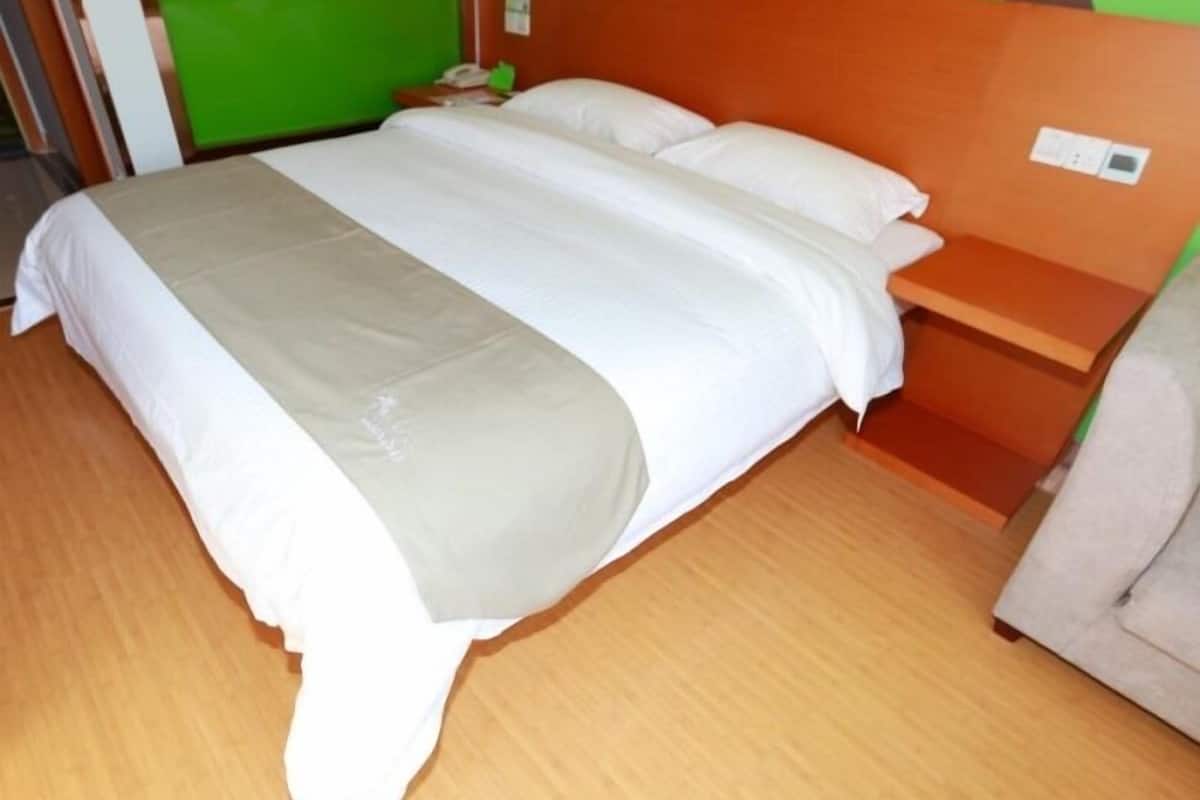 Ein ordentlich bezogenes Bett mit weißen Laken und einer grauen Decke, ein Nachttisch aus Holz mit einem Telefon und eine grüne Wand.