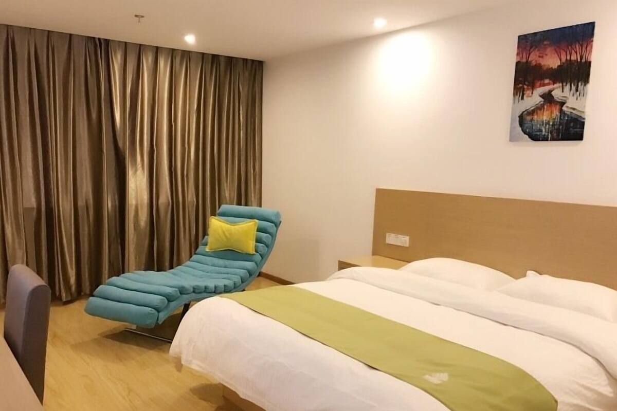 A hotel room with a bed, a blue reclining chair, a yellow pillow, and a painting on the wall.