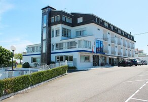 Un hotel de varios pisos con un llamativo letrero 'HOTEL', un balcón en el segundo piso y un estacionamiento al frente.