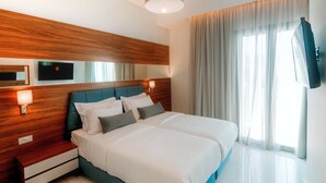 Quarto de hotel moderno com cama grande, cabeceira de madeira, abajures, televisão de tela plana e uma janela ampla com cortinas.