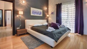 Ein modernes Schlafzimmer mit einem großen Bett, Nachttischlampen und Blick auf eine Stadtlandschaft durch transparente Vorhänge.