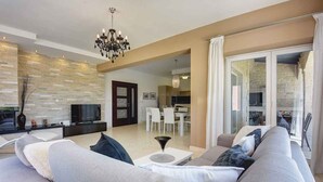 Ein modernes Wohnzimmer mit einer Couch, einem Fernseher und einer Deckenleuchte. Durch eine offene Tür ist eine Essecke mit Tisch und Stühlen zu sehen.
