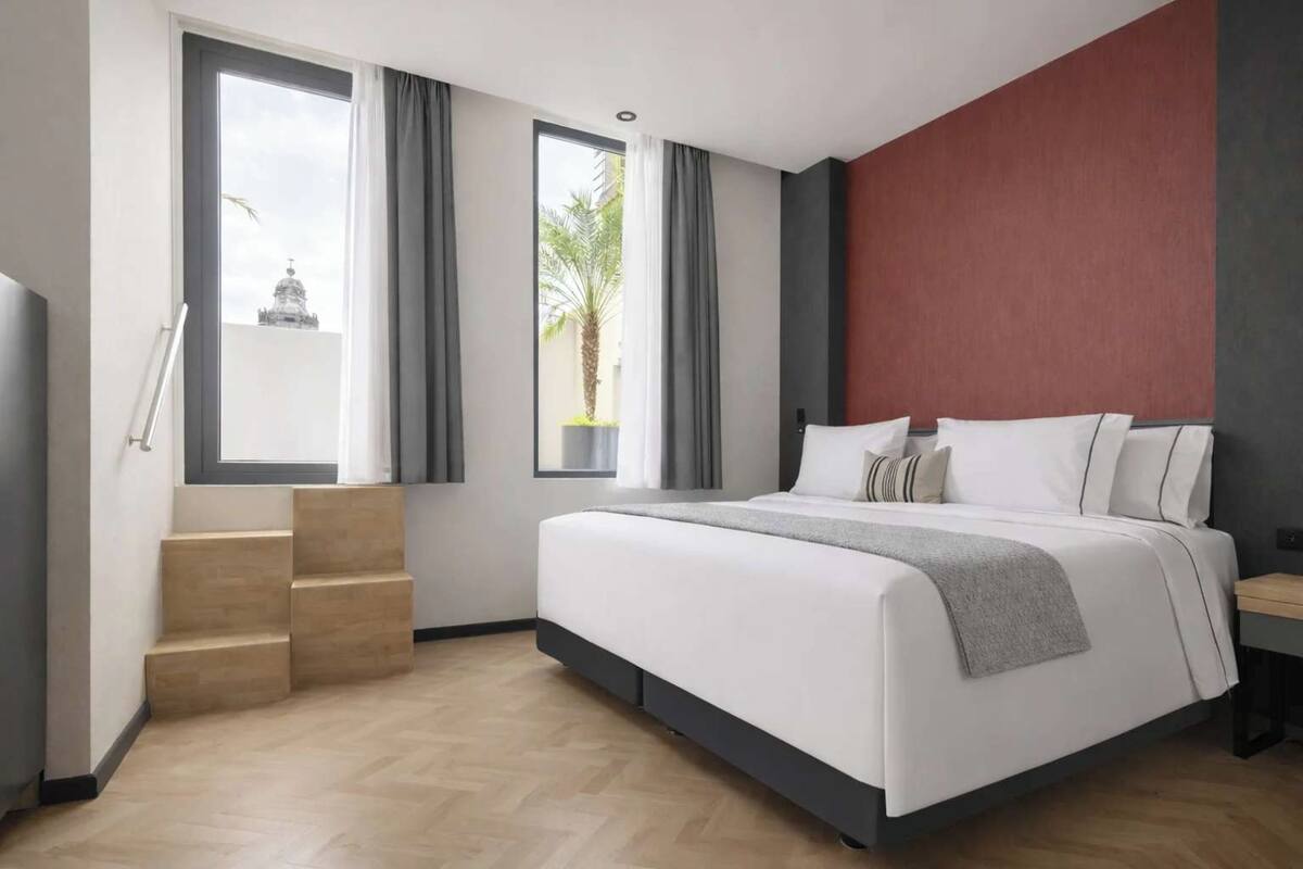 A modern hotel room with a bed, a small desk, a window with curtains, and a staircase.