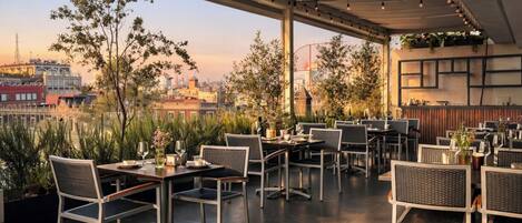 Un área de comedor al aire libre con mesas y sillas, vista del paisaje urbano y una noria al fondo.