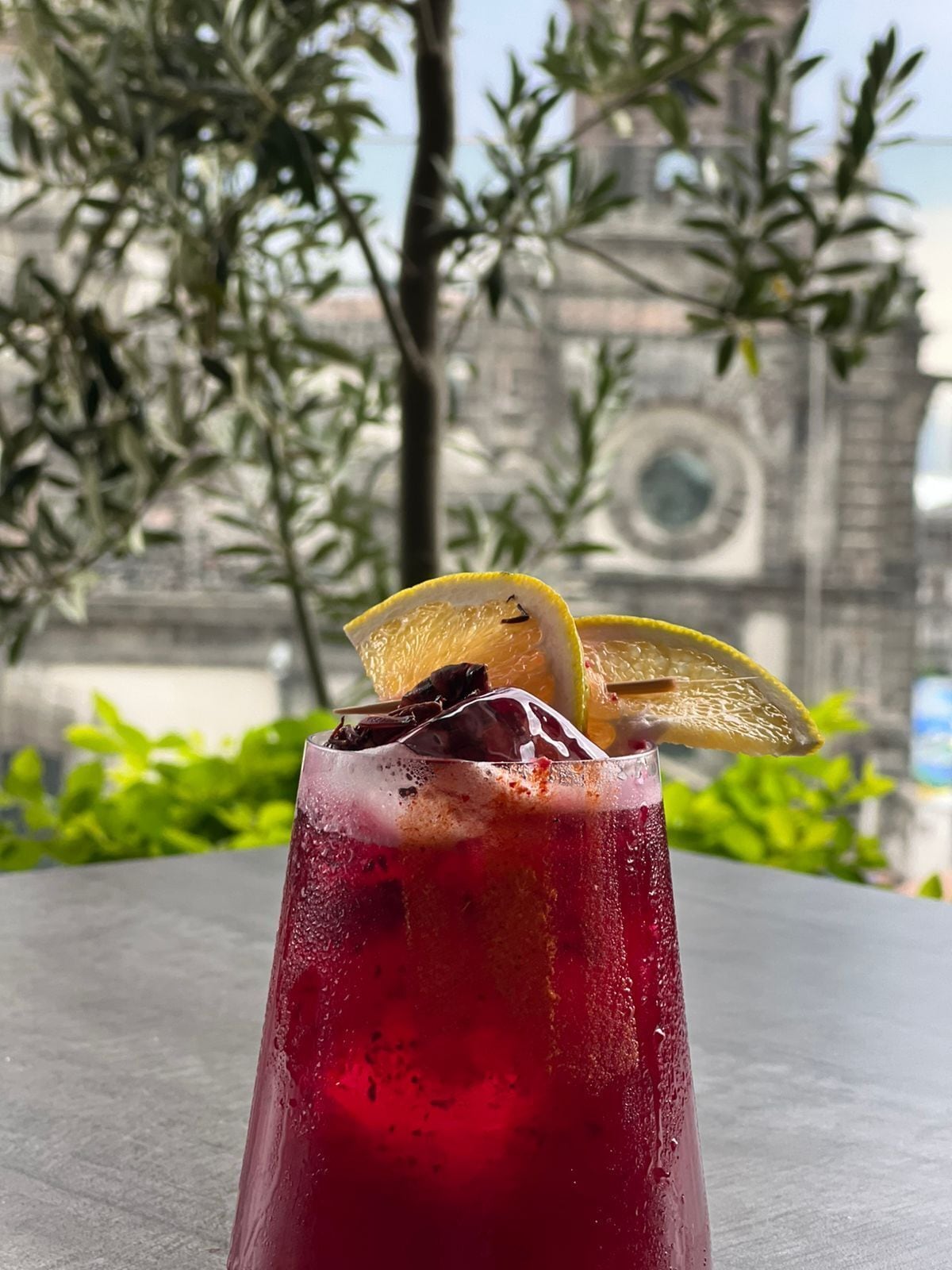 Un vaso alto con una bebida roja, una rodaja de limón y una fruta oscura en el borde, sobre una mesa con un fondo desenfocado de follaje y un edificio.