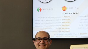 Um homem segurando folhetos de hotel, com uma tela exibindo preços e nomes de quartos de hotel ao fundo.
