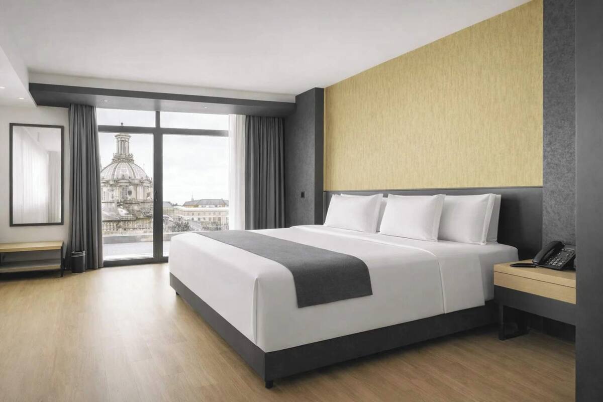 A modern hotel room with a large bed, a desk with a phone, and a view of a cityscape through a large window.