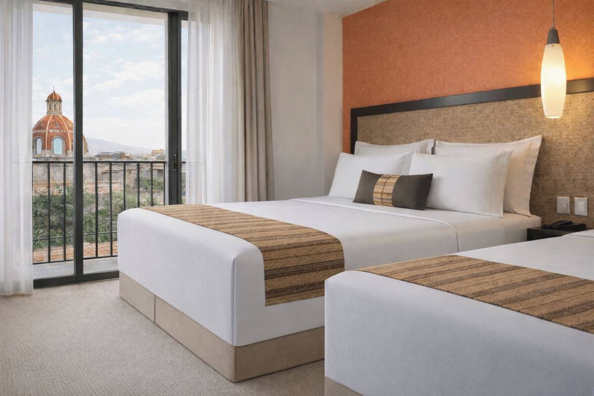 A hotel room with two beds, a view of a building through a balcony, and a hanging light fixture.