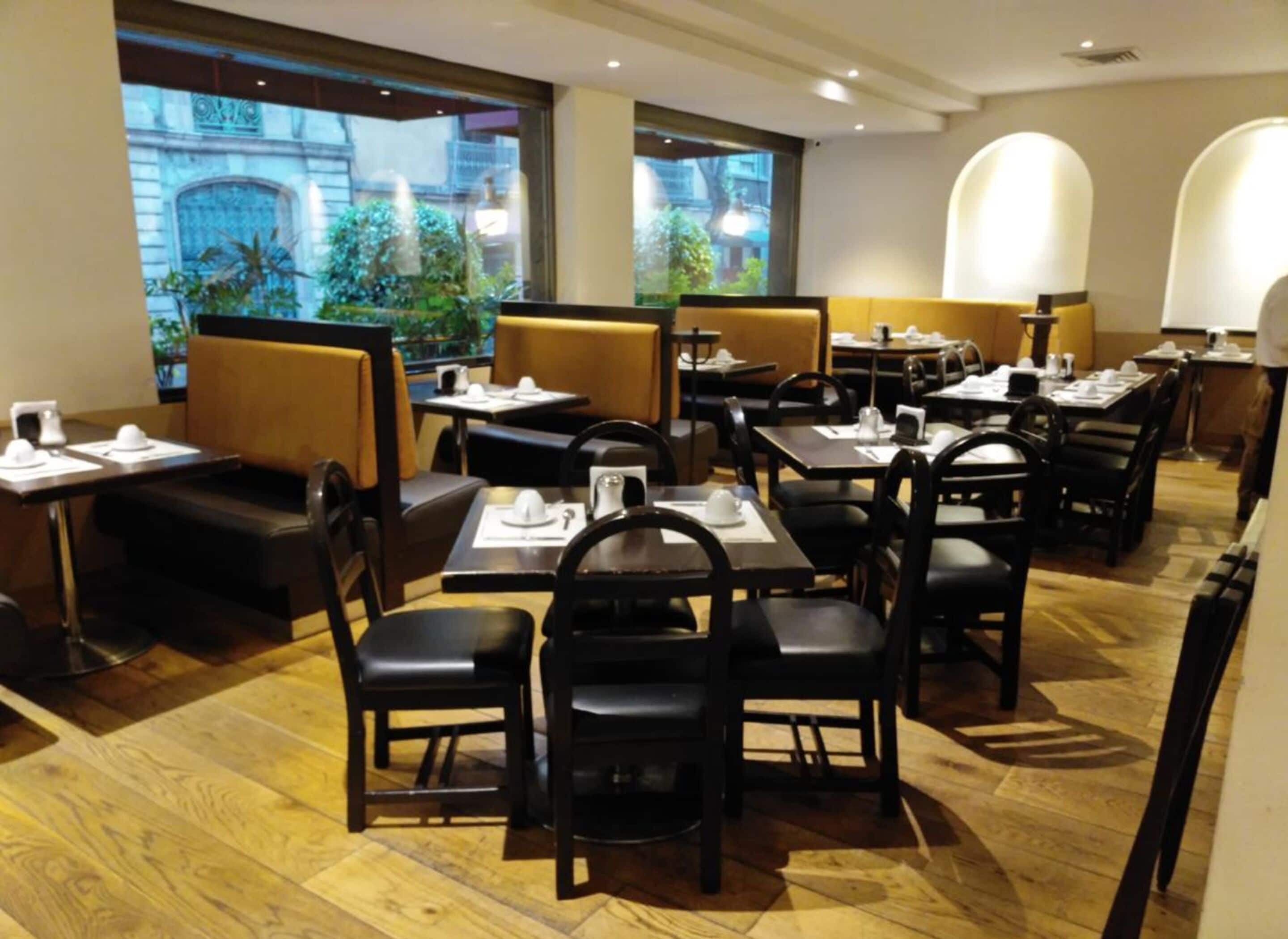 Un restaurante con cabinas y mesas, piso de madera y amplias ventanas con vistas a edificios y zonas verdes.