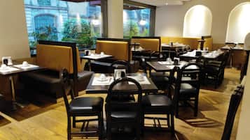 Un restaurante con cabinas y mesas, piso de madera y amplias ventanas con vistas a edificios y zonas verdes.