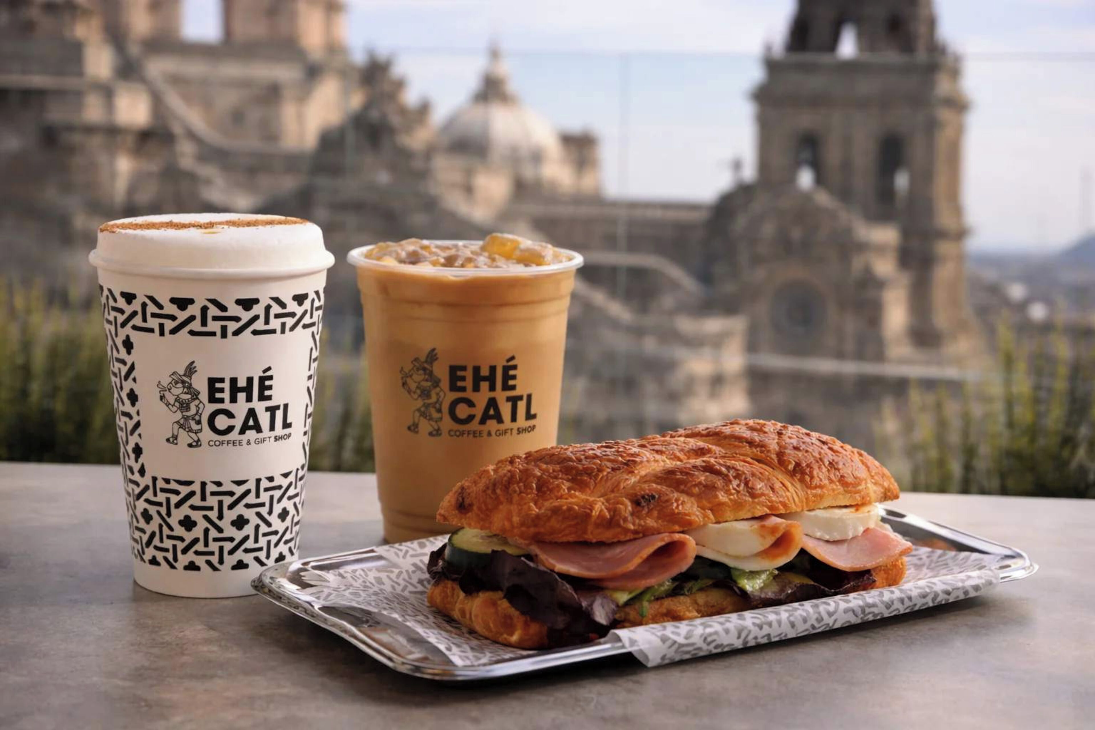 A sandwich, a coffee cup, and a pastry on a table with a cityscape in the background.