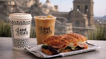 Un sándwich, una taza de café y un pastel sobre una mesa, con un paisaje urbano de fondo.