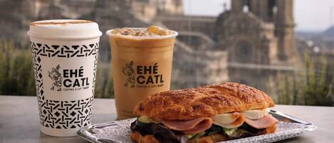 A sandwich, a coffee cup, and a pastry on a table with a cityscape in the background.